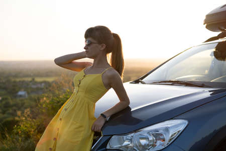 Happy young woman in yellow dress standing near her vehicle looking at sunset view of summer nature. Traveling and vacation concept.の写真素材
