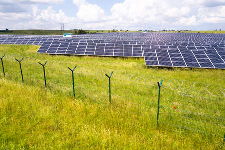 Aerial view of solar power plant on green field with protective wire fence around it. Electric...