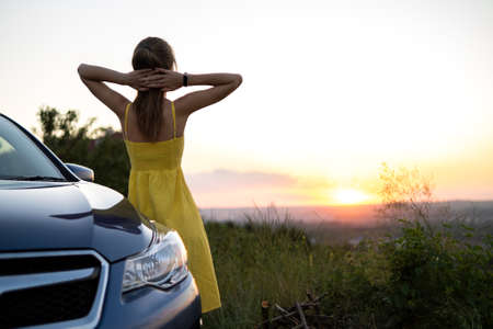 Happy young woman driver in yellow dress enjoying warm summer evening standing beside her car. Traveling and vacation concept.の写真素材