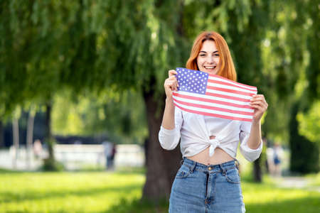 Happy young woman posing with USA national flag outdoors in summer park. Pretty girl celebrating...