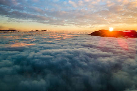 Aerial view of vibrant sunrise over white dense clouds with distant dark mountains on horizon.