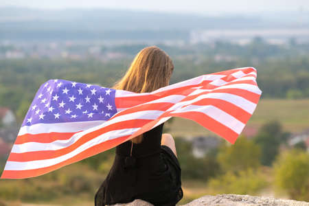 Young happy american woman with long hair holding waving on wind USA national flag on her sholders relaxing outdoors enjoying warm summer day.の写真素材