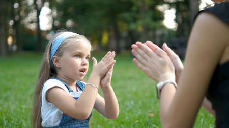 Pretty little child girl playing game with her mom clapping their hands together outdoors in green summer park.の写真素材