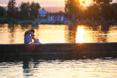 Side view of lonely woman sitting alone on lake shore on warm evening. Solitude and relaxing in...
