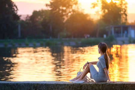 Back view of lonely young woman sitting alone on lake shore enjoying warm evening. Wellbeing and relaxing in nature concept.の写真素材