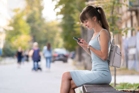 Young pretty woman messaging on mobile phone on warm summer day sitting on a city street bench outdoors.の写真素材
