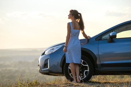 Happy woman driver in blue summer dress enjoying warm evening near her car. Travel and vacations concept.の写真素材
