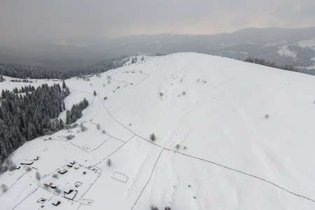 Aerial winter landscape with small rural houses between snow covered forest in cold mountains.