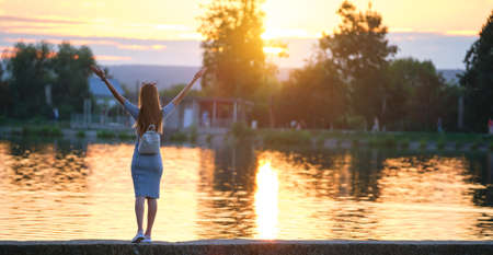 Rear view of successful woman standing alone on lake shore with raised up hands on warm evening. Success and meditation concept.の写真素材