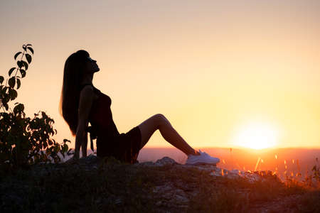 Young slim woman in black short dress sitting on a rock relaxing outdoors at summer sunset. Fashionable female enjoying warm evening in nature.の写真素材