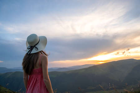 Young woman in red dress standing on grassy field on a windy evening in autumn mountains enjoying...