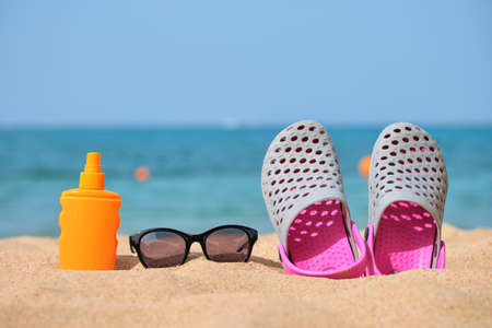 Closeup of clogs shoes, sunscreen and black protective sunglasses on sandy beach at tropical seaside...