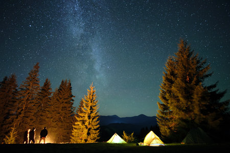 Silhouette of hikers resting besides burning bonfire near illuminated tourist tents on camping site in dark mountains under night sky with sparkling stars. Active lifestyle and outdoor living conceptの写真素材