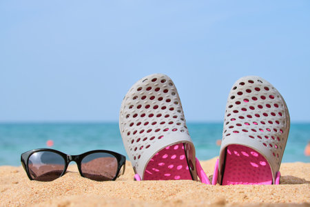 Closeup of clogs shoes and black protective sunglasses on sandy beach at tropical seaside on warm...