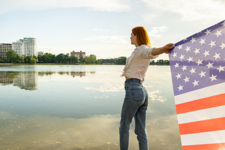 Red haired girl holding USA national flag in her hands. Positive young woman celebrating United States independence day.の写真素材