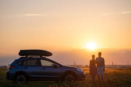 Happy couple standing near their car at sunset. Young man and woman enjoying time together travelling by vehicleの写真素材