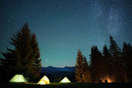 Silhouette of hikers resting besides burning bonfire near illuminated tourist tents on camping site in dark mountains under night sky with sparkling stars. Active lifestyle and outdoor living conceptの写真素材