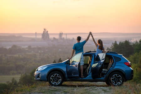 Happy couple spending time together near their SUV car during honeymoon road trip at warm summer evening. Young man and woman enjoying road trip travelling by vehicle in natureの写真素材