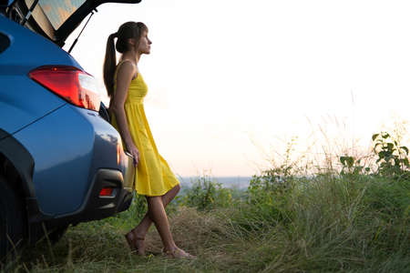 Happy young woman in yellow dress standing near her vehicle looking at sunset view of summer nature. Travelling and vacation conceptの写真素材