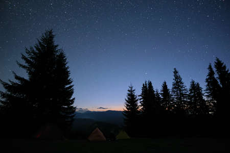 Brightly illuminated camping tents glowing on campsite in dark mountains under night stars covered sky. Active lifestyle and traveling conceptの写真素材