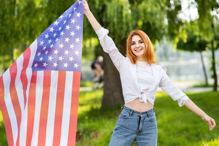 Happy young red haired woman posing with USA national flag standing outdoors in summer park....