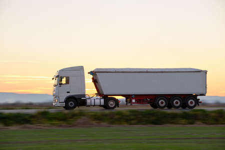 Semi-truck with tipping cargo trailer transporting sand from quarry driving on highway hauling goods in evening. Delivery transportation and logistics conceptの写真素材