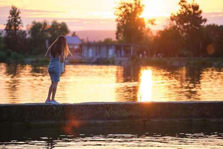 Rear view of lonely woman standing alone on lake shore on warm evening. Solitude and relaxing in nature conceptの写真素材