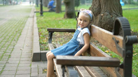 Small happy child girl sitting on a bench resting in summer parkの写真素材