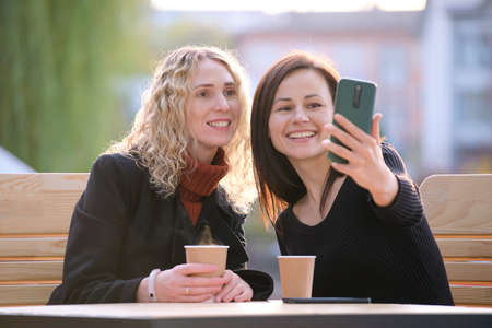 Two happy women making selfie with mobile phone sitting at city street cafe. Female friends enjoying time together outdoorsの写真素材