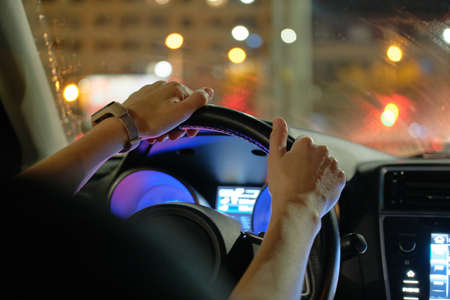 Close up of driver hands holding steering wheel driving car with blurred city street lights on...