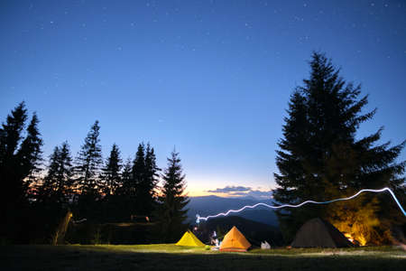 Brightly illuminated camping tents glowing on campsite in dark mountains under night stars covered...