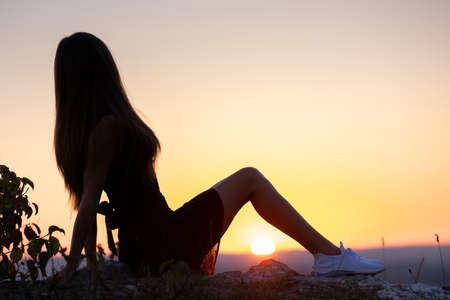 Young elegant woman in black short dress sitting on a rock relaxing outdoors at summer evening. Fashionable female enjoying warm sunset in natureの写真素材