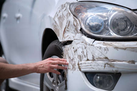 Driver hand examining dented car with damaged fender parked on city street side. Road safety and vehicle insurance conceptの写真素材