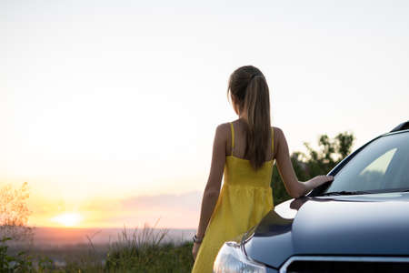 Happy young woman in yellow dress standing near her vehicle looking at sunset view of summer nature. Travelling and vacation conceptの写真素材