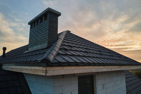 Chimney on house roof top covered with metallic shingles under construction. Tiled covering of...