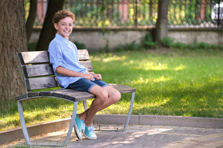 Young happy child boy relaxing sitting on bench in summer park. Positive kid enjoying summertime outdoors. Child wellbeing conceptの写真素材