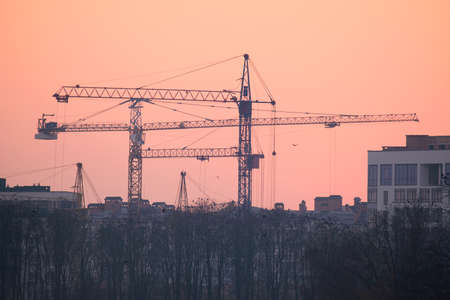 Dark silhouette of tower cranes at high residential apartment buildings construction site at sunset. Real estate developmentの写真素材