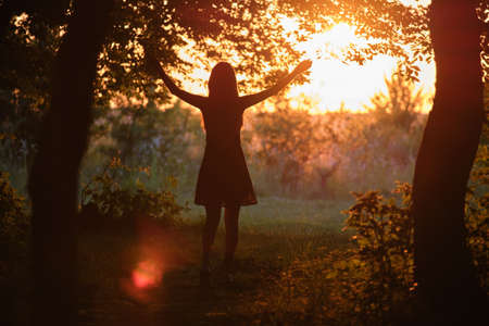 Dark silhouette of young woman walking alone through dark forest joyfully raising her hands in...