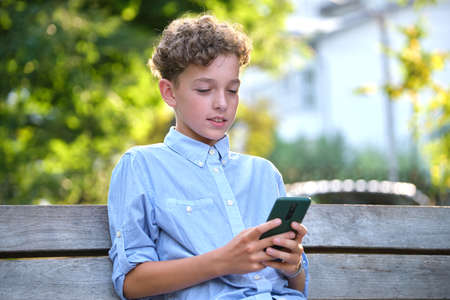 Young boy playing game on his smartphone outdoors in summer park. Addiction from electronic gadgets...