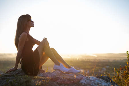 Young slim woman in black short dress sitting on a rock relaxing outdoors at summer sunset. Fashionable female enjoying warm evening in natureの写真素材