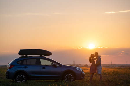 Happy couple spending time together near their SUV car during honeymoon road trip at warm summer evening. Young man and woman enjoying road trip travelling by vehicle in natureの写真素材