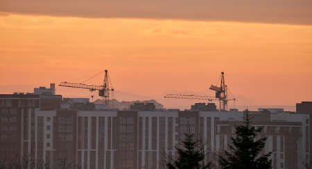 Dark silhouette of tower cranes at high residential apartment buildings construction site at sunset. Real estate developmentの写真素材