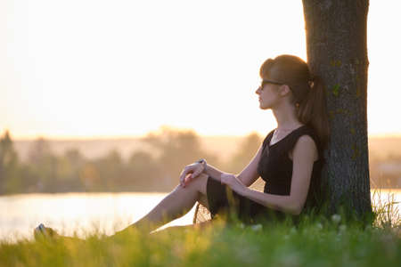 Lonely woman sitting alone on green grass lawn leaning to tree trunk on lake shore on warm evening. Solitude and relaxing in nature conceptの写真素材