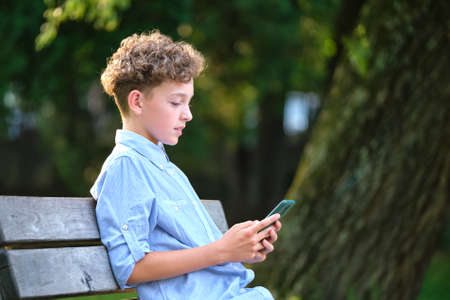 Young boy browsing his smartphone outdoors in summer park. Using cellphone for leasure and education conceptの写真素材