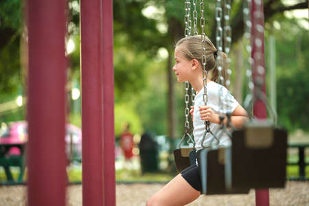 Young pretty happy smiling teenage small girl having fun on playground swings on summer vacations sunny dayの写真素材