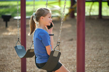 Happy young child girl talking on cellphone sitting outdoors on swings in summer park