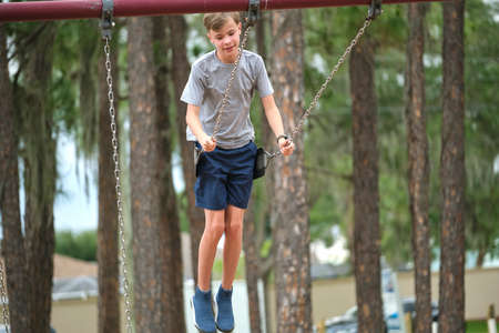 Young handsome teenage boy playing alone on playground swings on summer vacations sunny day