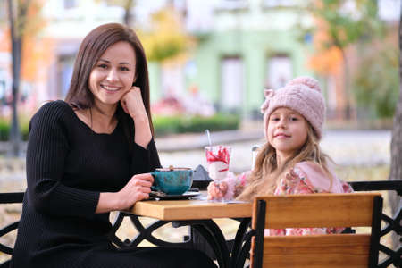 Young pretty woman having good time with her child daughter sitting at street cafe with hot drinks...