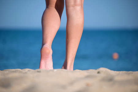 Close up of female feet walking barefoot on white grainy sand of golden beach on blue ocean water backgroundの写真素材