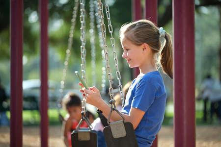 Happy child girl browsing her mobile phone sitting on swing in park during summer vacationsの写真素材